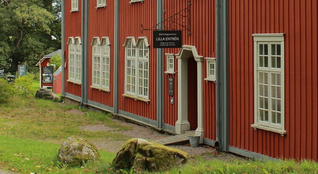 Stor byggnad med stående rödmålad träpanel och mångspröjsade fönster med en utstickande butiksskylt ovanför en dörröppning. 