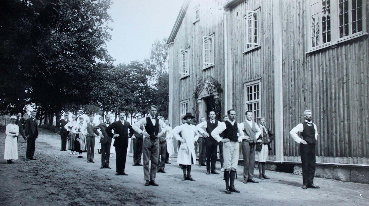 Ett tjugotal personer i gammaldags kläder står i strikta led med händerna i midjan framför ett stort trähus. Fotot är svartvitt och från 1915 ca