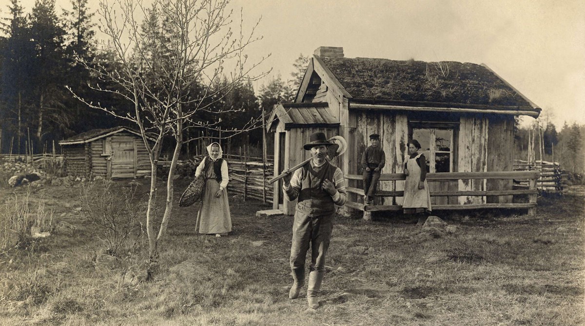 Svartvitt foto föreställande en man med stövlar, hatt och väst och en spade över axeln med en äldre kvinna med huckle och två barn framför ett mycket litet bostadshus med torvtak. 