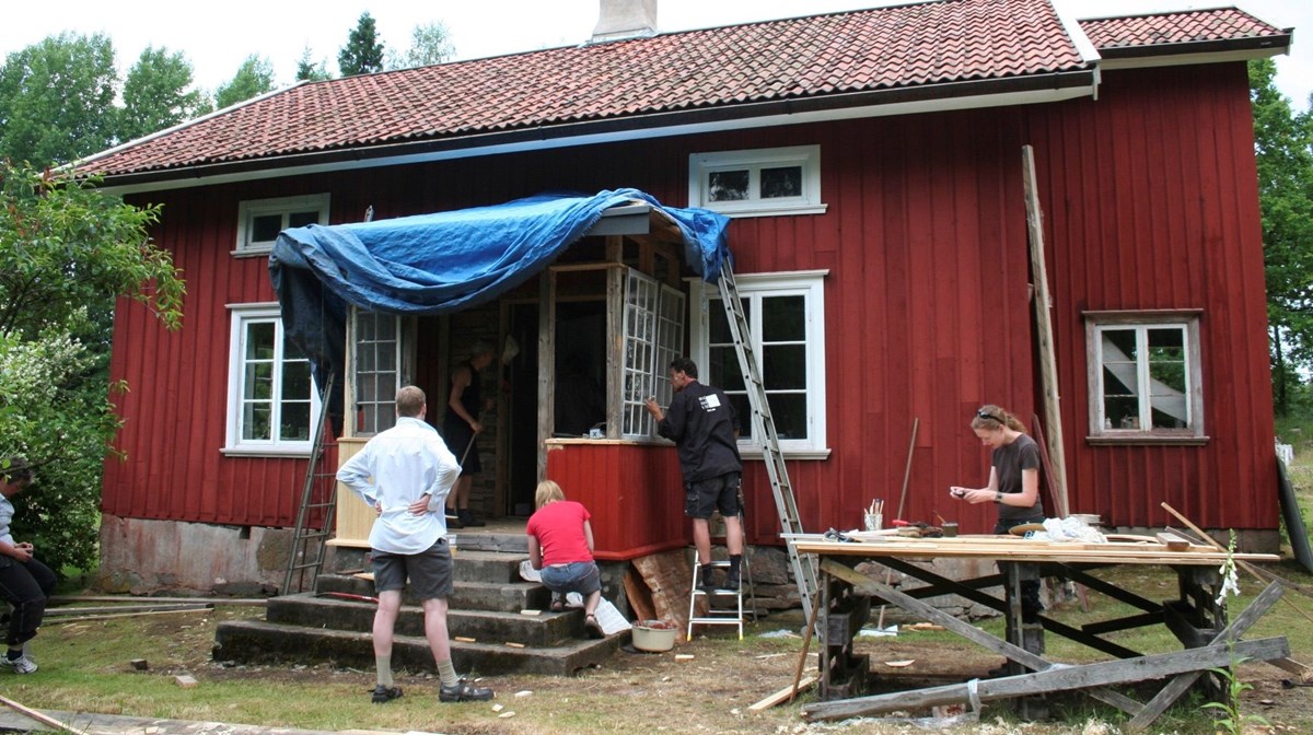 Ett rött trähus med personer som bygger på en glasveranda.