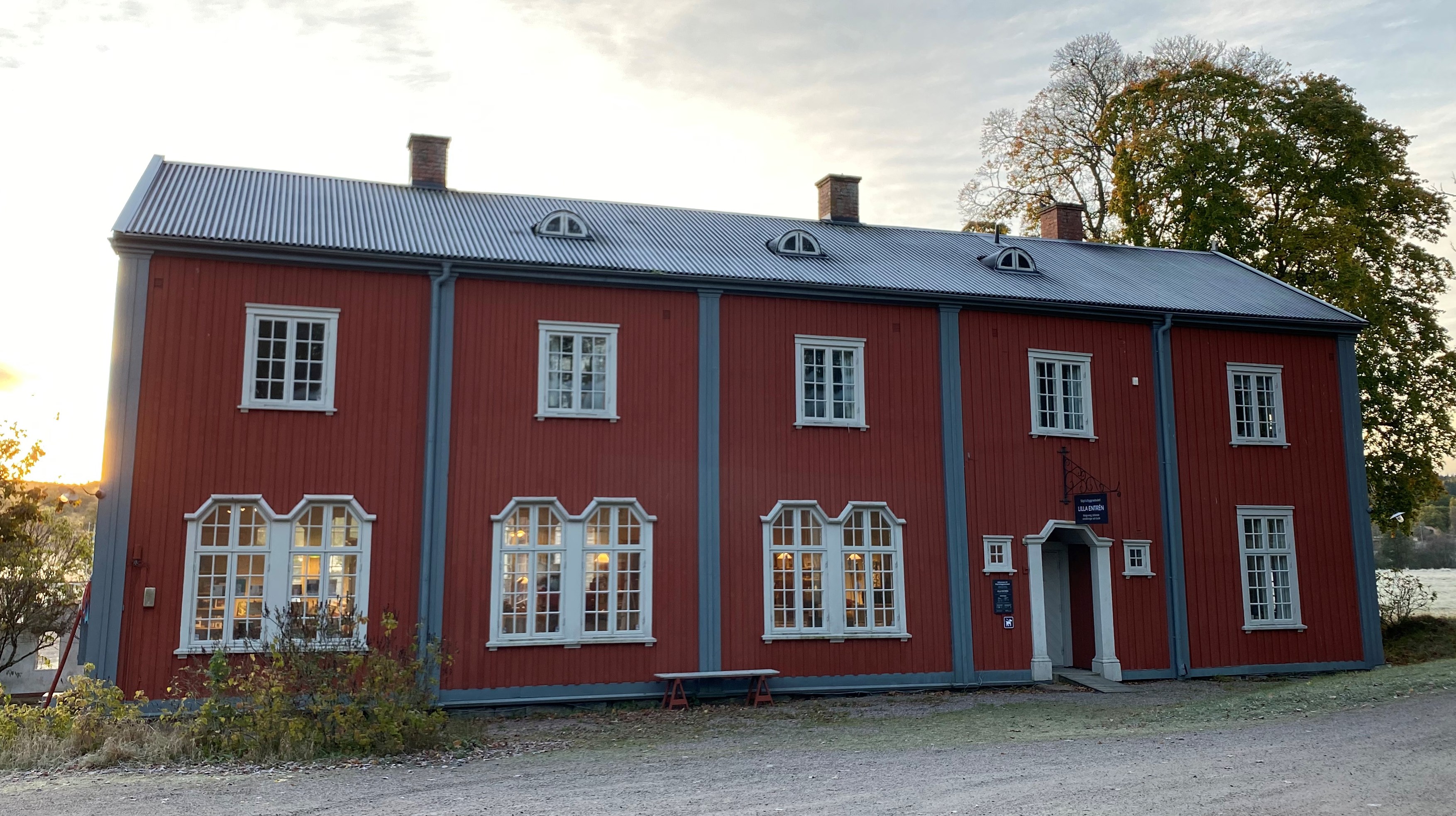 Ett stort rött trähus med grå och ljusgrå fönster och lister. I bakgrunden gröna träd. Taket är svart och med rimfrost. En låg sol lyser bakom huset.