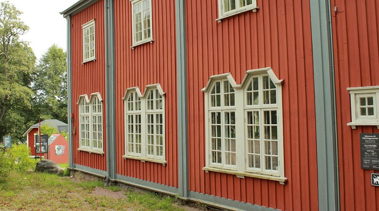 Detalj av rödbrunt trähus med blågrå lister och ljusgrå fönster. Fönstren på nedervåningen är stora och småspröjsade. På övervåningen enklare fönster.