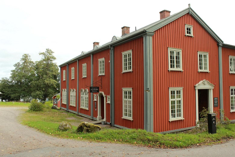 Ett stort rödbrunt hus med gråblå detaljer och ljusgrå fönster. Huset syns med två fasader med två entréer i ljusgrått. I förgrunden grusad väg och grästäckt vägren