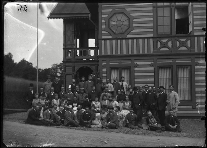En grupp med människor står framför ett hus med randig fasad. Svartvitt foto från ca 1900.
