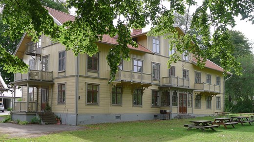Ett stort gulbrunt hus med grå foder och röda fönster. I förgrunden lövade trädgrenar
