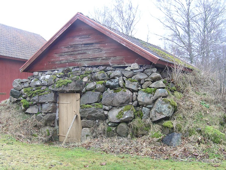 En stor jordkällare med mosstäckta stenar och ett rödmålat litet hus uppepå. Dörren är stöttad med en bräda