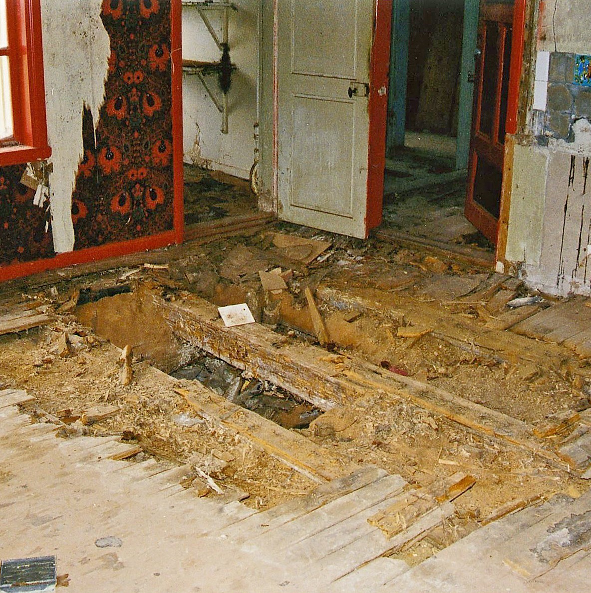 En interiör med uppbrutet trägolv med tydliga skador och frilagd sågspånsisolering. Väggarna har flagnande tapeter.