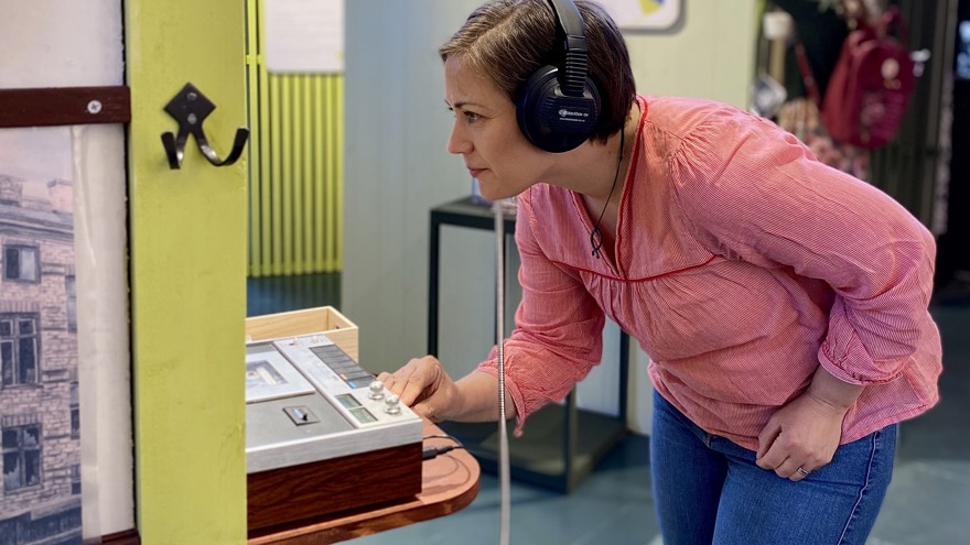 En kvinna med stora hörlurar står lutad framför en gammal kassettbandspelare och verkar läsa på något.