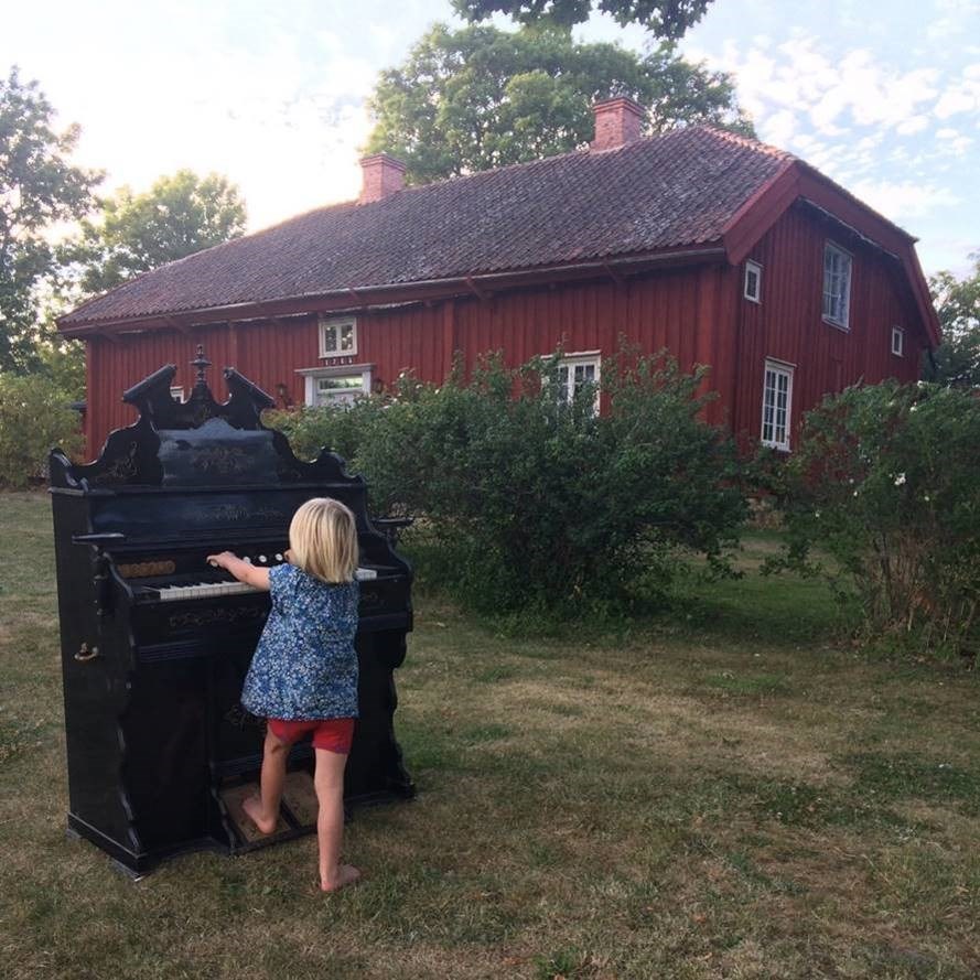 En sommarkväll med ett barn som spelar på en svartmålad gammal orgel på en gräsmatta. I fonden ett rödmålat 1700-talshus