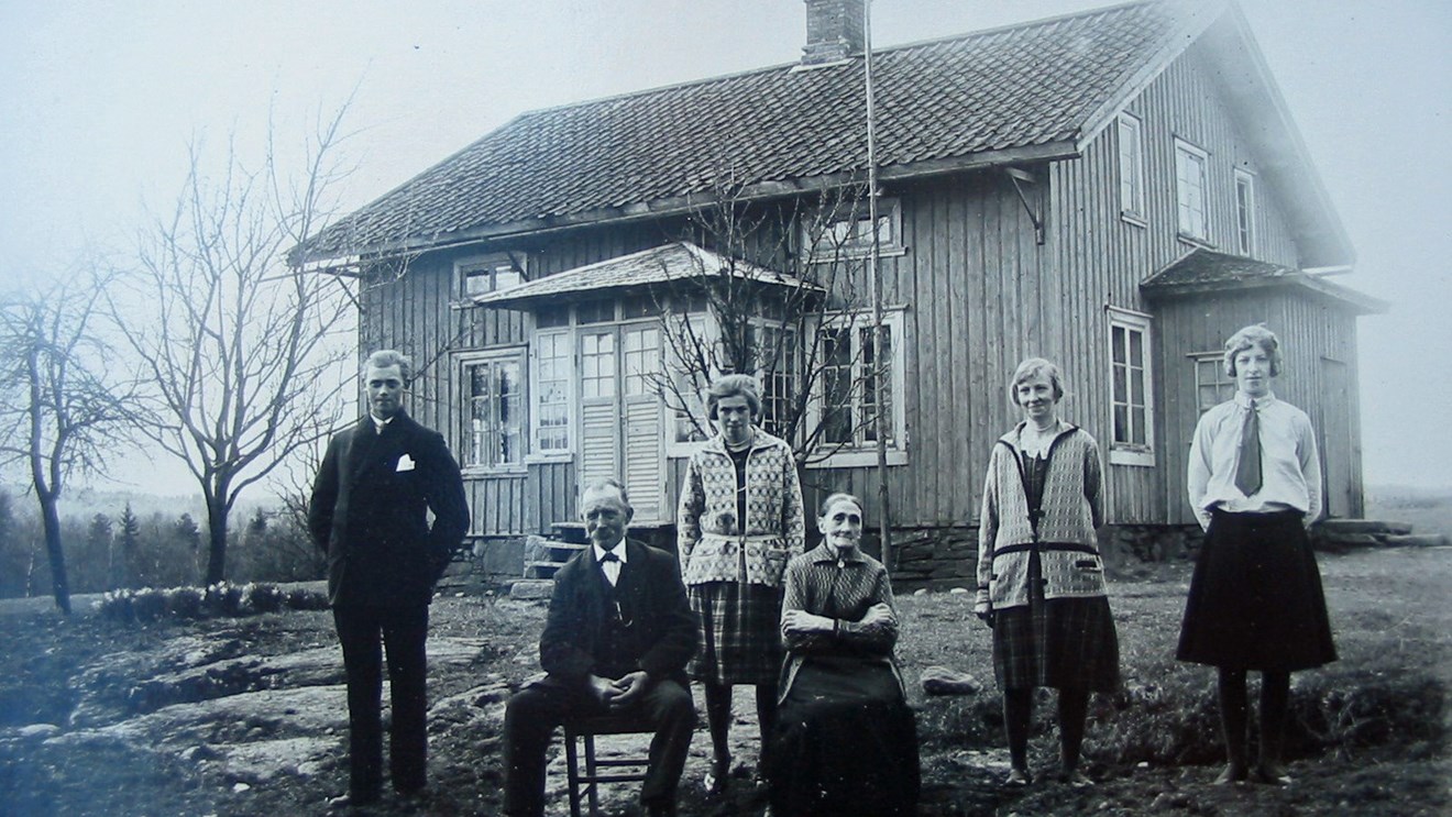Foto från ca 1900 i svartvitt med sex personer framför ett hus i trä med liten glasveranda