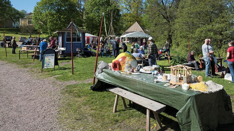 utomhus i solen bord med produkter och folk som pratar med varandra. I bakgrunden träd och ett gult hus