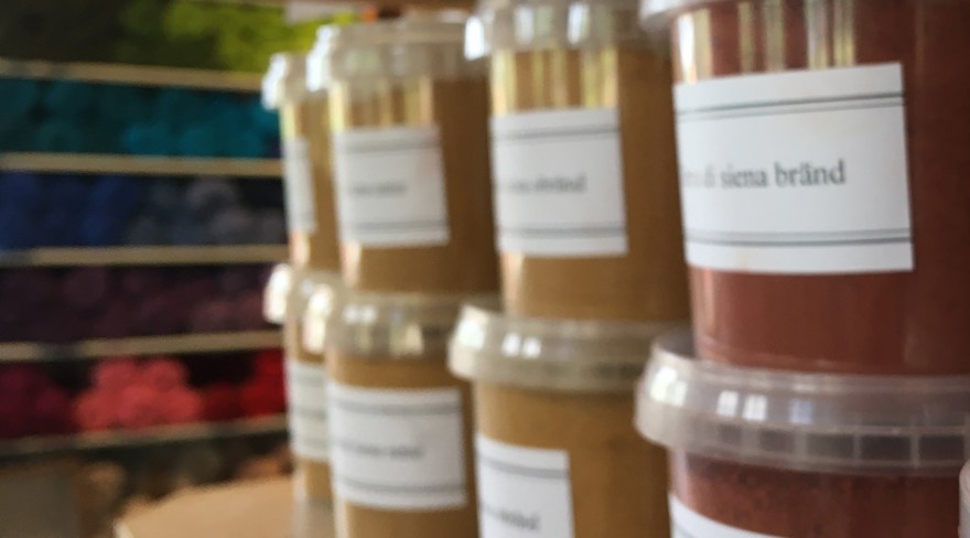 Burkar med pigment står på en hylla inomhus. I bakgrunden ull i flera färger i en hylla.