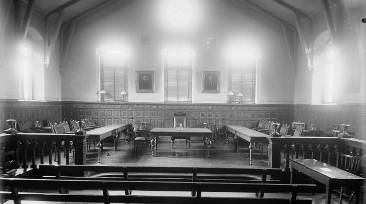 Svartvitt foto från 1930 som visar en stor sal med högt i tak, väggarnas nedre del är täckt med mörk träpanel. I mitten står ett bord med en hög stol - detta flankeras av två bord med flera stolar. I förgrunden är ett skrank och närmast i bild träbänkar. Bilden visar en tingssal från 1930.