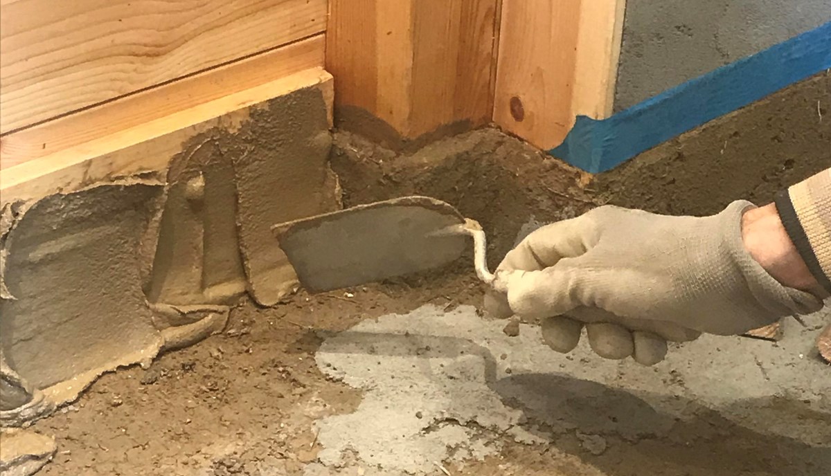 en hand håller en murslev och murar med lera i skarven mellan trävägg och gjutet golv