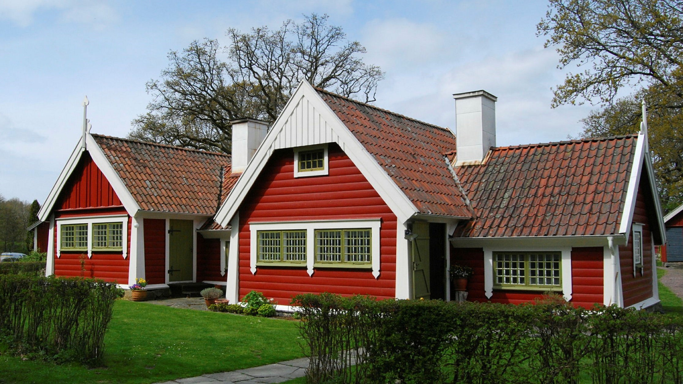 Ett hus med flera huskroppar och stort takfall målat i rött med vita foder och gröna fönster.
