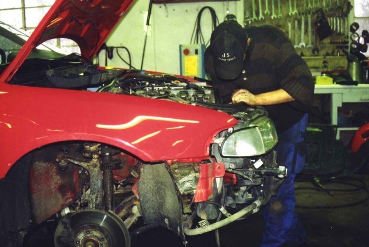 En person med keps står och mekar i motorhuven på en röd söndermonterad bil