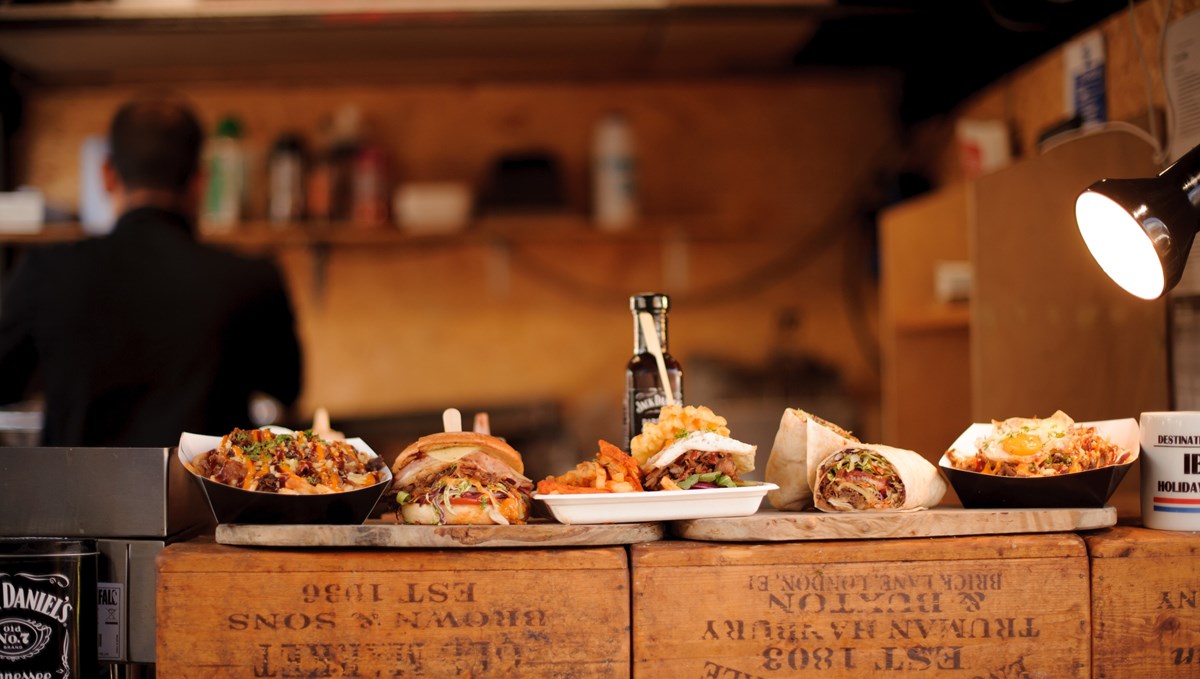 Streetfood-rätter upplagda på trälådor, i bakgrunden en person i ett kök.