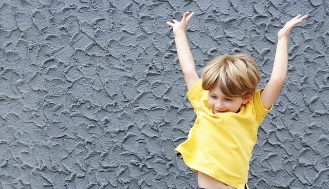 Ett barn i gul tröja och ljust hår ser glad ut och håller armarna över huvudet. Blå bakgrund