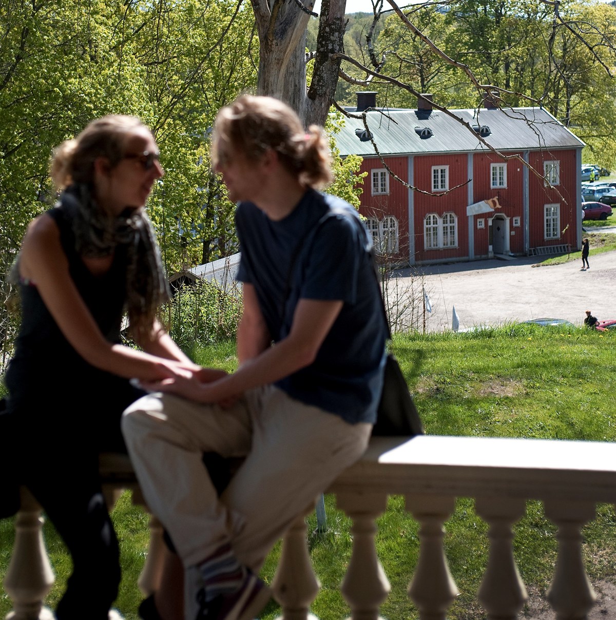 två personer sitter på ett verandaräcke och med rött hus i bakgrunden
