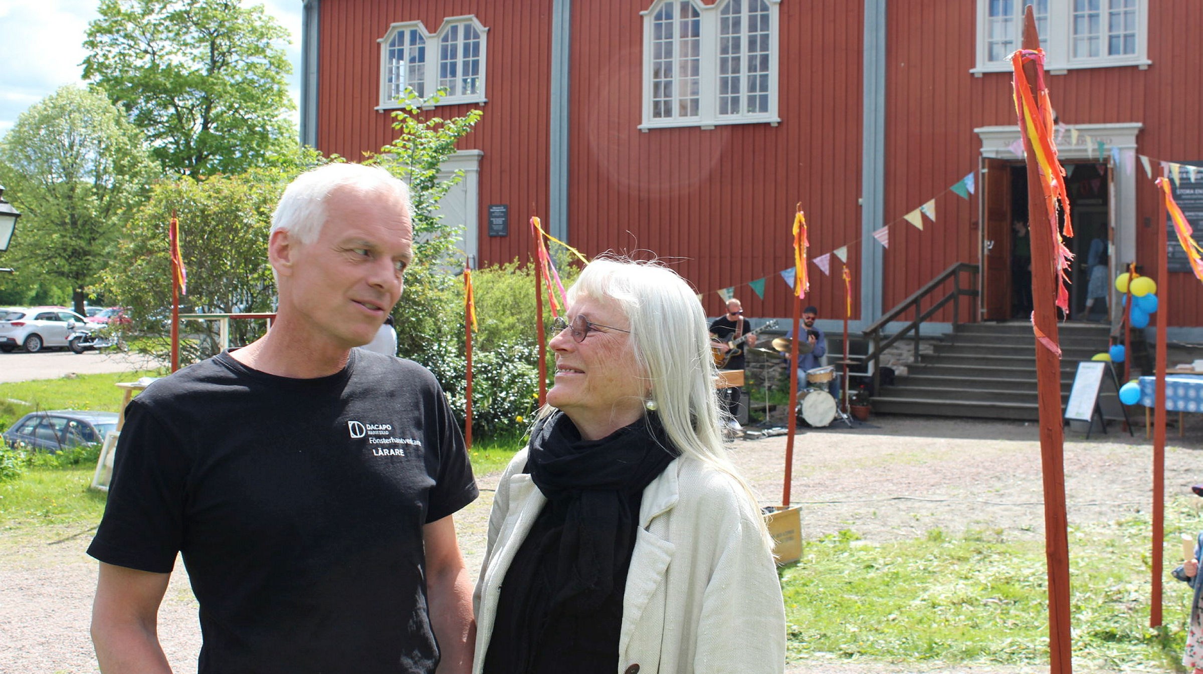 Två personer ler mot varandra, i bakgrunden ett stort rött hus och i gräskanten är rödmålade stolpar med tygbitar fästa