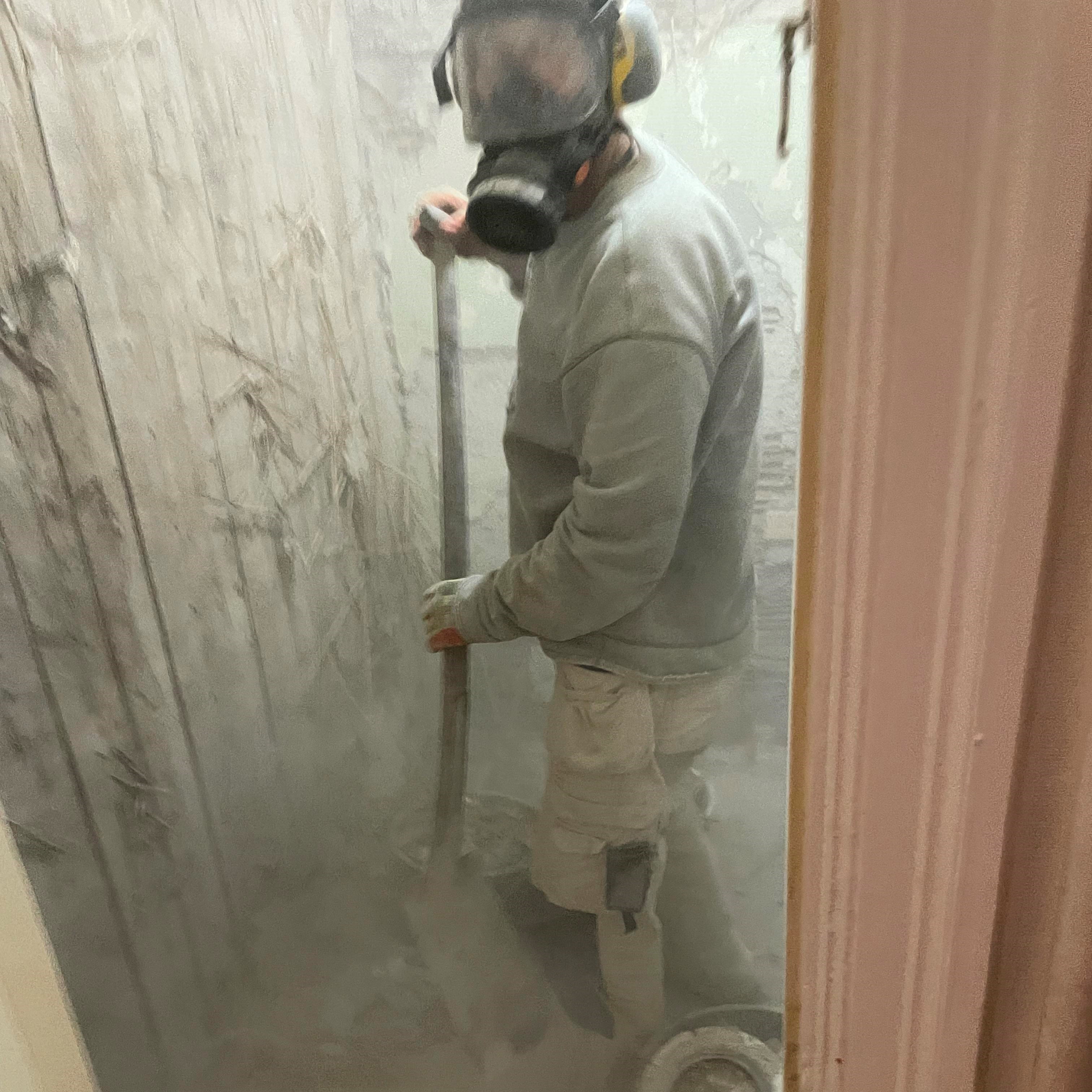 Person med skyddskläder och andningsmask står med en spade i ett litet rum som är grått av byggdamm som flyger i luften.