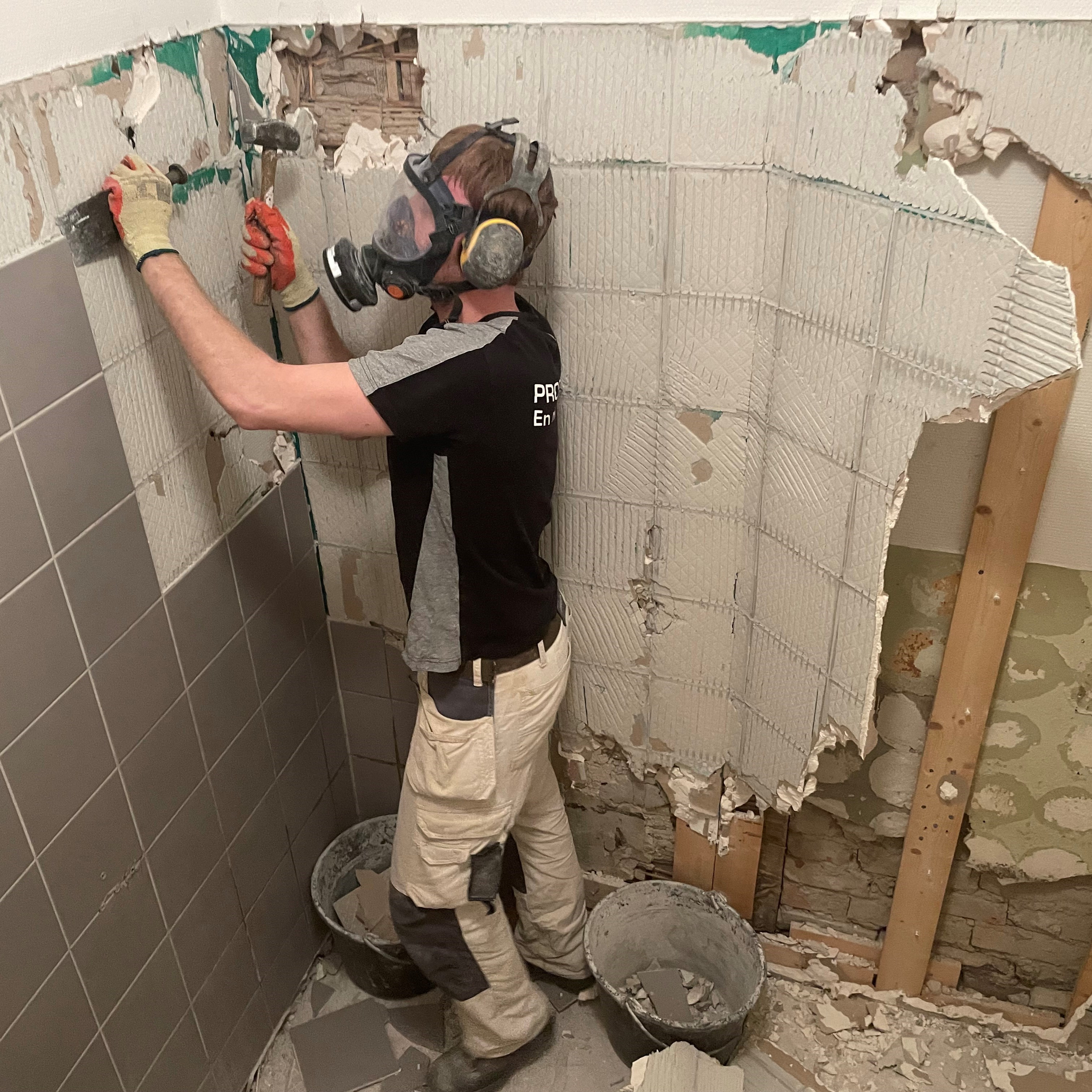 Hantverkare med andningsmask och handskar knackar bort kakel från en vägg. 