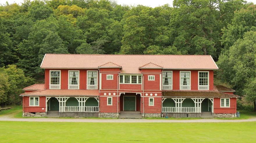 Ett stort rött trähus med rött tegeltak och vita detaljer. Flera entréer med gröna dörrar. I bakgrunden högvuxna lövträd och i förgrunden gräsmatta.