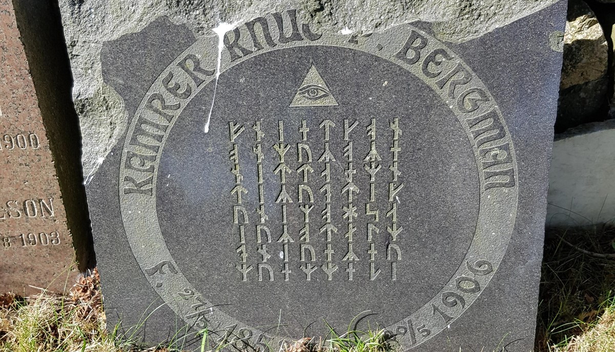 Kvadratisk sten med inskriptioner i runor och svenska språket - det är en gravsten från 1906 över Kamrer Knut Bergman. Stenen står lutad mot en mur omgiven av andra äldre stenar.