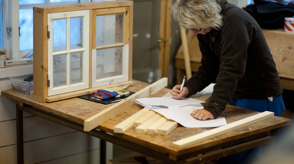 Inomhus ett bord med ett fönster i karm stående och en person som skriver på ett papper. det ligger virke och verktyg på bordet