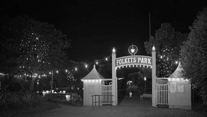 Svartvit bild visar nattbild med upplyst entré med grindar och skylt med text Folkets park. Träd med belysning i.