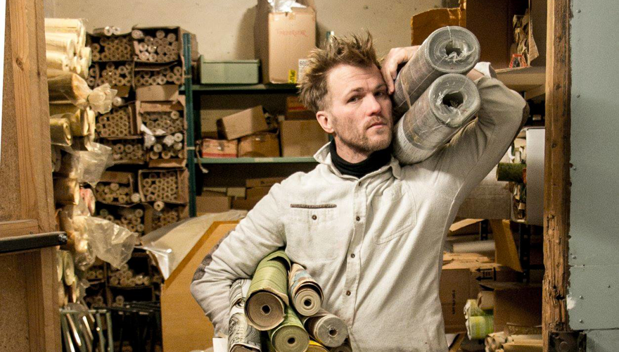 En man står framför fulla lagerhyllor och håller stora tapetrullar över axeln.