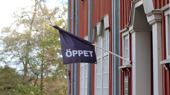 En flagga med texten öppet hänger utanför en entré på en rödmålad träbyggnad med dekorativt utformade snickeridetaljer.