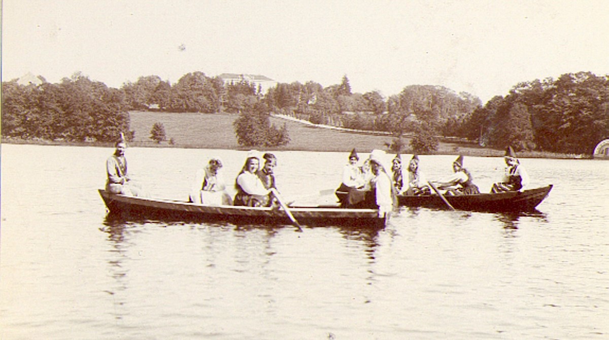 Svartvitt gammalt foto med två båtar fulla av kvinnor i folkdräkt. I bakgrunden tronar en herrgård