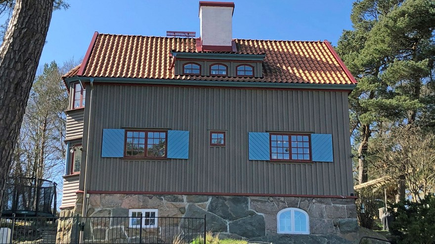 Ett grågrönt trähus sett nedifrån. Huset har rött tegeltak, röda fönster och blå fönsterluckor. Skorstenen är vit.