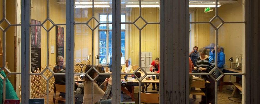 Innanför spröjsade fönster ses ett tiotal personer sitter och står runt ett bord