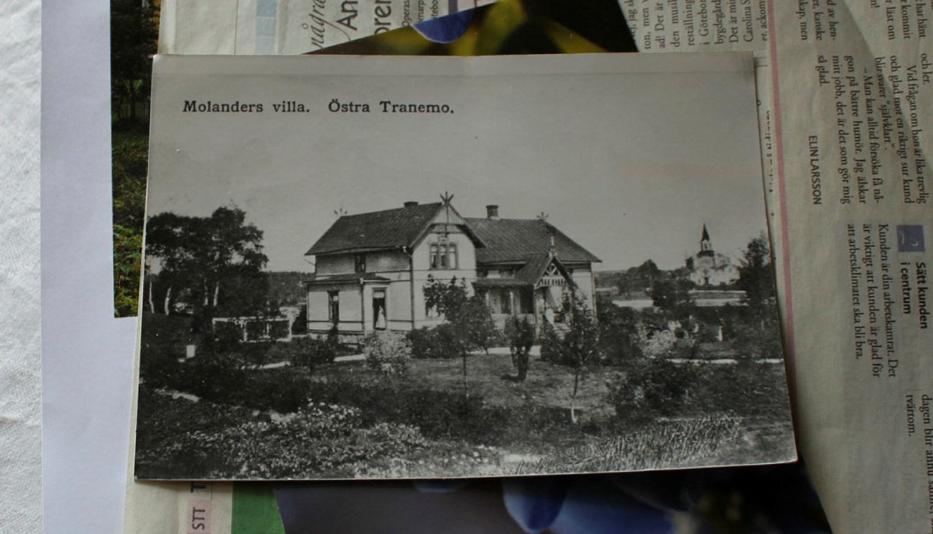 Ett svartvitt vykort över ett äldre hus ligger på ett bord med tidningar under