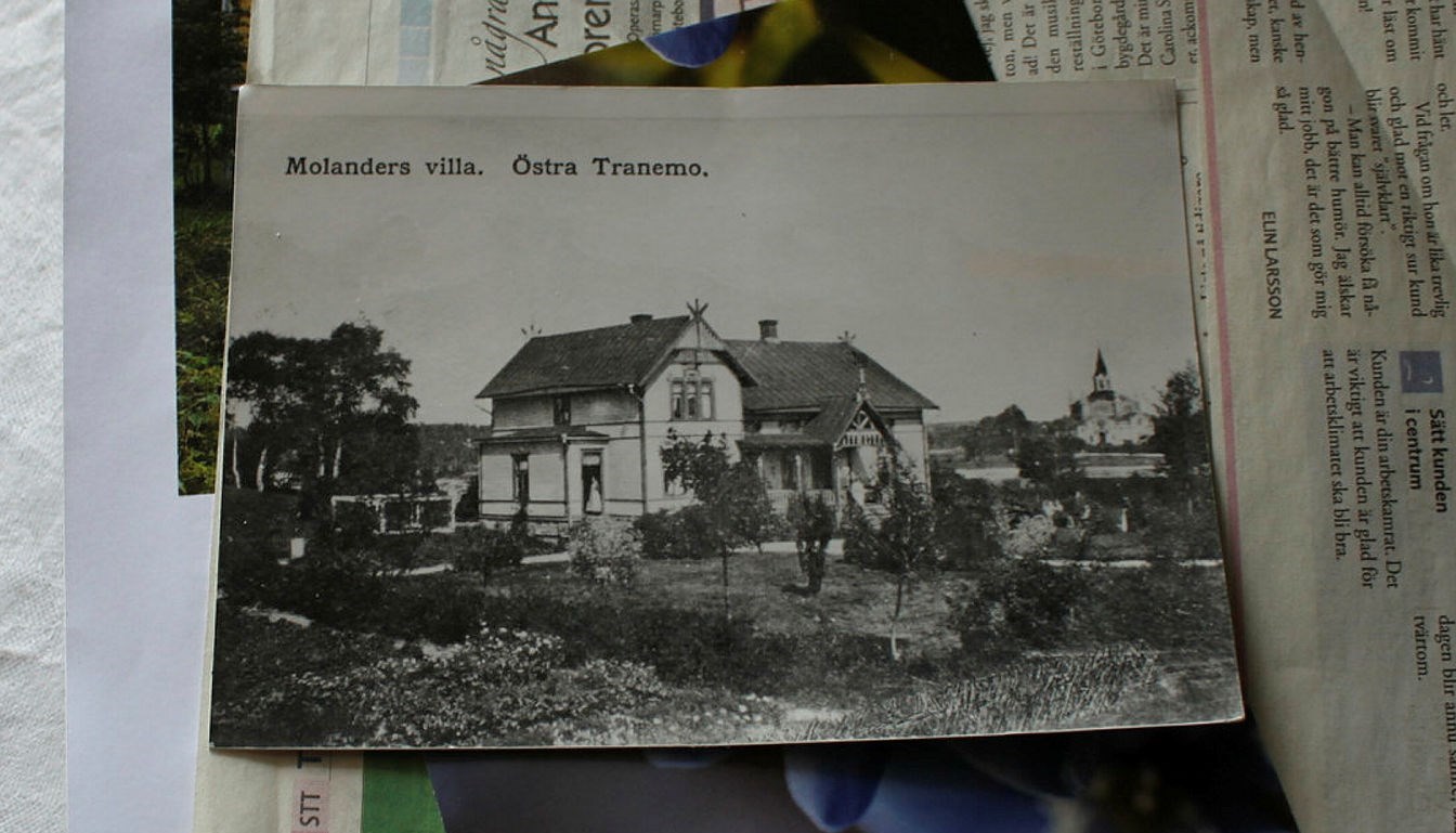 Ett svartvitt vykort över ett äldre hus ligger på ett bord med tidningar under 
