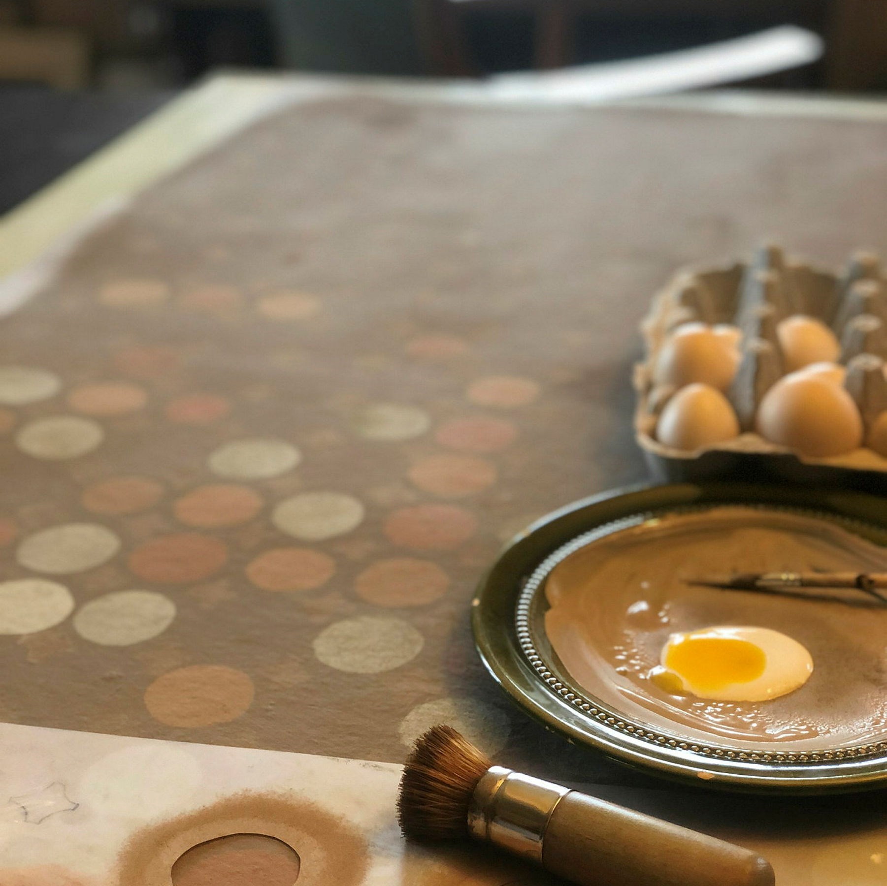 målning på ett bord med runda prickar i olika bruna och beiga kulörer och bredvid ligger ägg och en pensel