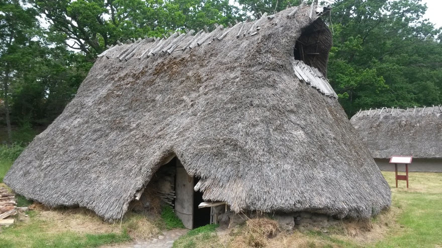 Stort forntidshus täckt med vasstak och med en ingångsöppning nedtill och en öppning i gaveln högst upp. Grönskande omgivning med träd och ett liknande hus som skymtar bakom