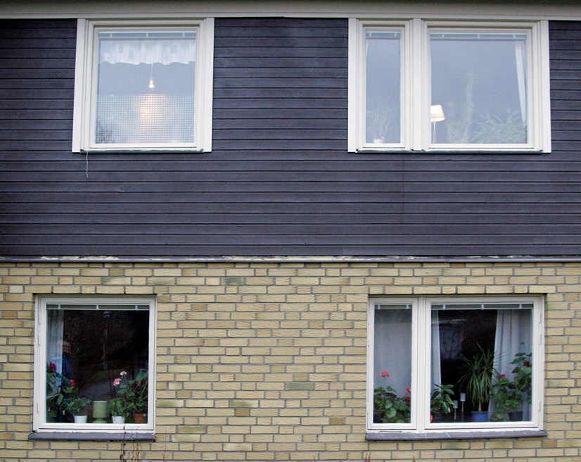 Detalj av ett hus med bottenvåningen i gult tegel och övervåningen i liggande brunmålad panel. Fönster är vita.