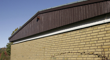 Gavelparti på gult tegelhus. Den övre delen av gaveln är klädd med stående brunmålad träpanel.