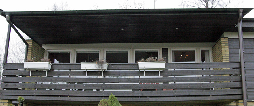 Detalj av balkongen över entrén. Räcket är brunmålad liggande glespanel. Taket över balkongen är en utdragen del av husets tak, vilande på stolpar i balkongens hörn. Rummet innanför balkongen har fem vitmålade fönster och en vitmålad balkongdörr.