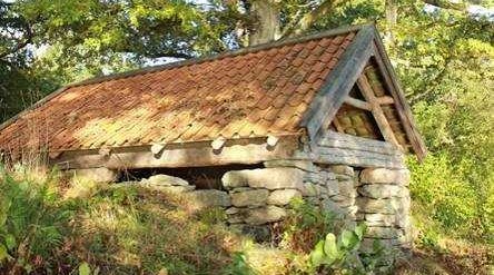 En liten byggnad i sten och trä med tegeltak, delvis dold i vegetationen.