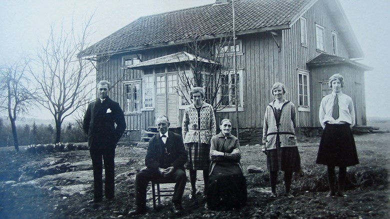 Foto från 1920 ca där fyra personer står och två sitter framför ett hus med glasveranda, omgivningarna visar landsbygd och skog i närheten