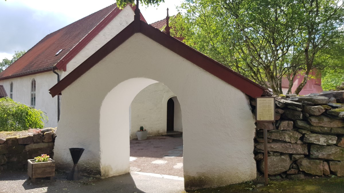 En murad och putsad öppning i en stenmur. Väggarna är vitmålade och taket har rödmålade vindskivor. I bakgrunden skymtar en vitmålad kyrka och högvuxna träd