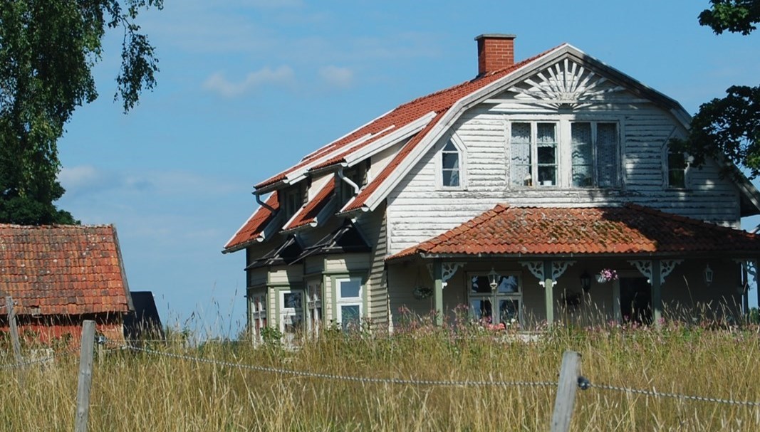 Ett stort vitt hus med rött tegel och snickarglädje står i solskenet. I förgrunden en äng och till vänster en liten rödmålad ekonomibyggnad