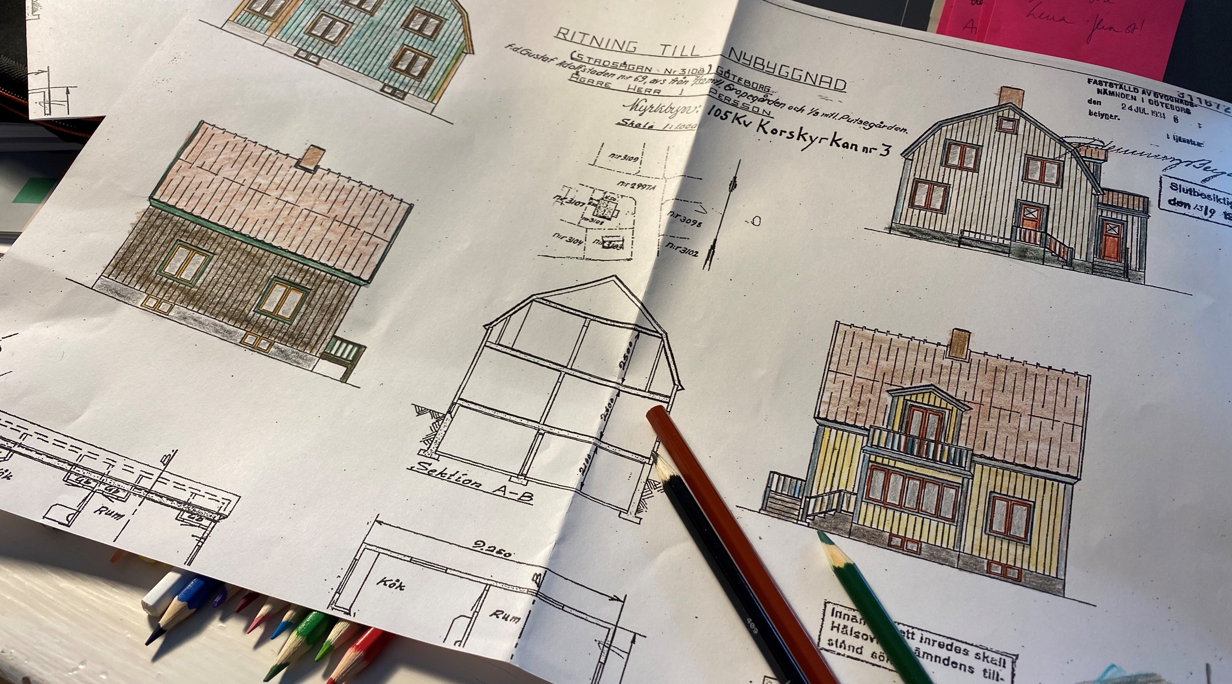 Ett papper med arkitektritning av ett hus och de olika fasaderna färglagda med olika kulörer. Under och på ritningen ligger färgpennor