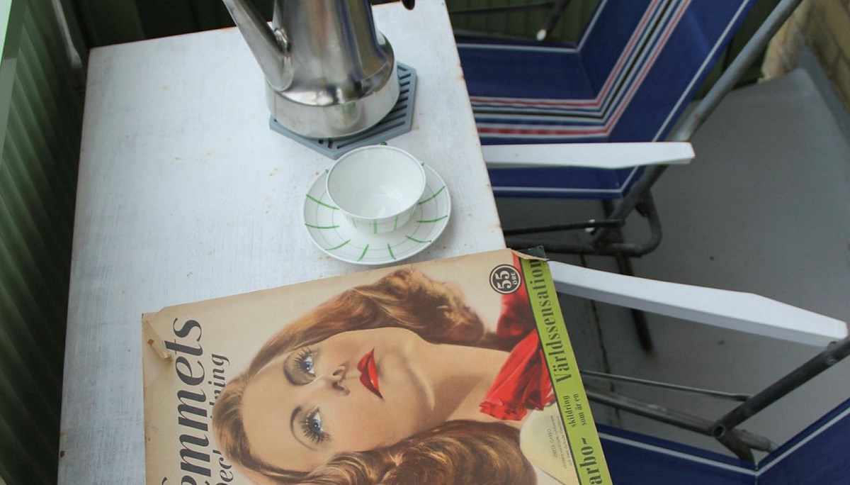 På en balkong två enkla hopfällbara stolar och ett bord som hänger på balkongräcket. På bordet en gammal Hemmets journal med tecknat kvinnoansikte på samt en kaffekopp på fat och en termos i metall.