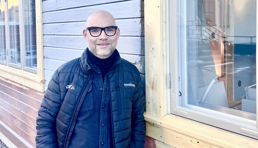 En leende man står lutad mot en husfasad med träpanel.