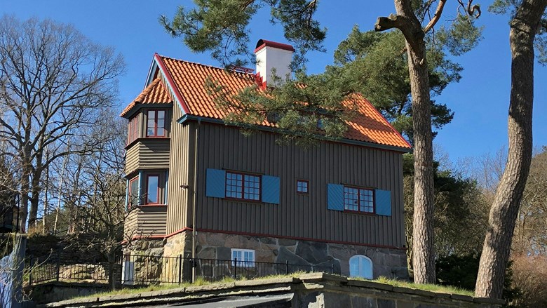 Ett stort gröngrått hus i trä med rött taktegel och röda fönster. Till vänster är två höga tallar och i bakgrunden lövträd